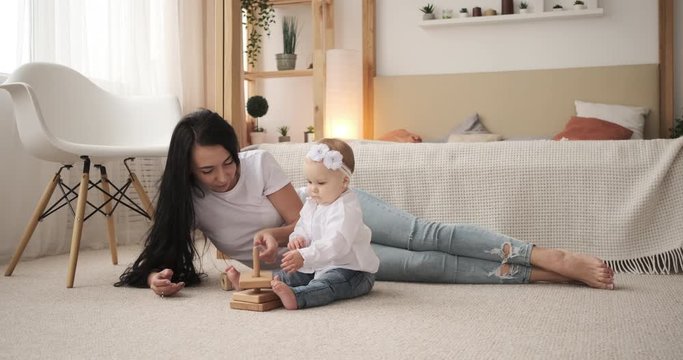 Mother and baby daughter playing with toy block lying on carpet in bedroom
