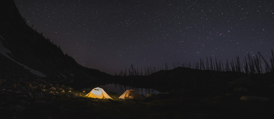 Lit tent next to an alpine lake with the star sky above