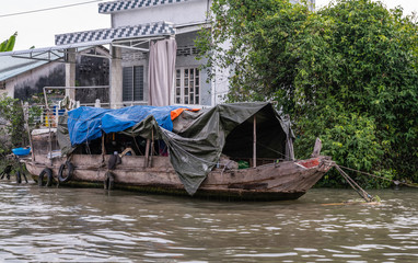Fototapeta na wymiar Cai Be, Mekong Delta, Vietnam - March 13, 2019: Along Kinh 28 canal. Blue and orange tarp covered old wooden sloop in front of white house and green foliage on brown water.