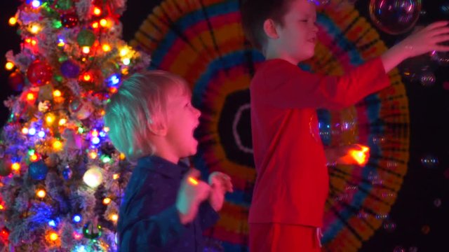 Soap bubbles fall on top of children. Boys catch them with candles in their hands against background of Christmas tree. Bright lights of garlands and decorations. Colors of rainbow. Shallow focus