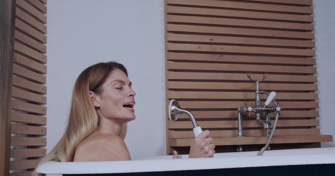 Beautiful young happy woman taking a bubble bath wahing herself singing a song with shower handset having fun doing healthy evening routine at home.