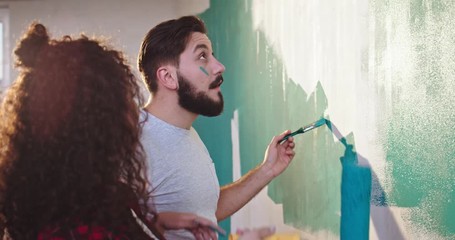 Cute and beautiful young Caucasian woman painting walls in the apartment in blue color with a wall brush roller and having fun with her husband, laughing and painting on each other's faces. - Powered by Adobe