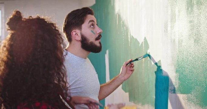 Cute and beautiful young Caucasian woman painting walls in the apartment in blue color with a wall brush roller and having fun with her husband, laughing and painting on each other's faces.