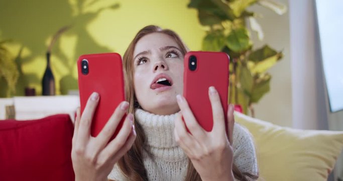 Funny young red-haired woman using two smartphones showing stupid cray face grimaces at camera laughing indoors. Home people. Humor concept.