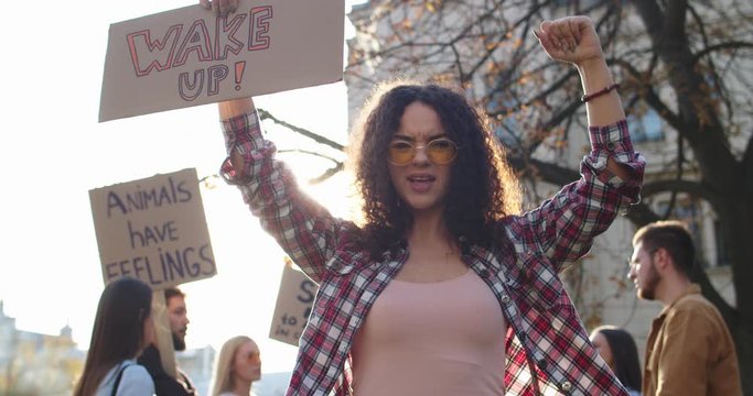 Young Caucasian curly pretty girl in sunglasses holding a poster "Wake up" and fighting for rights of animals and nature activism at the protest outside. Portrait shot.