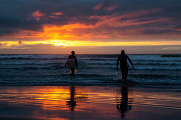 Fototapeta premium Surfers walking towards the Ocean on the sandy beach during a dramatic summer sunset. Taken in Seaside, Oregon, United States of America.