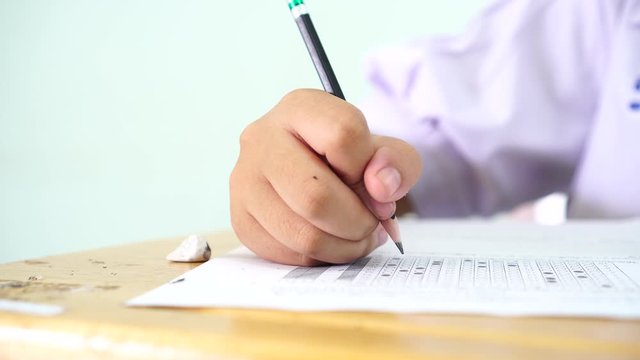 Teen young Asian students group concentrate one's mind writing test in exam on paper answer sheet and sitting on taking final examination ,in rows desk at classroom with Thai student uniform