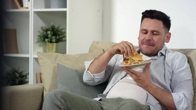 Middle aged Caucasian man holding plate and eating delicious burger while sitting on couch and watching interesting program on TV in the living room