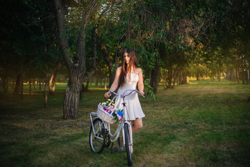 girl in a white skirt and a vest with a bicycle and a basket full of flowers in the park in the evening