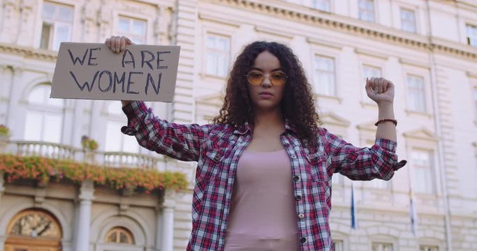 Beautiful young Caucasian woman with curly hair and in sunglasses fighting as the feminists activist at the female rights demonstration with a banner "We are women". Outdoors.