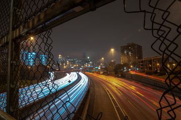 White and red light trails coming and going to downtown Chicago on a cloudy misty night viewed through an opening in a chain link fence guard rail on an overpass above highway or expressway at night.