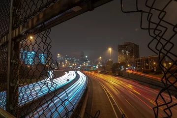 Poster White and red light trails coming and going to downtown Chicago on a cloudy misty night viewed through an opening in a chain link fence guard rail on an overpass above highway or expressway at night.
