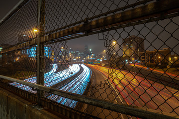 White and red light trails coming and going to downtown Chicago on a cloudy misty night viewed through an opening in a chain link fence guard rail on an overpass above highway or expressway at night. - Powered by Adobe