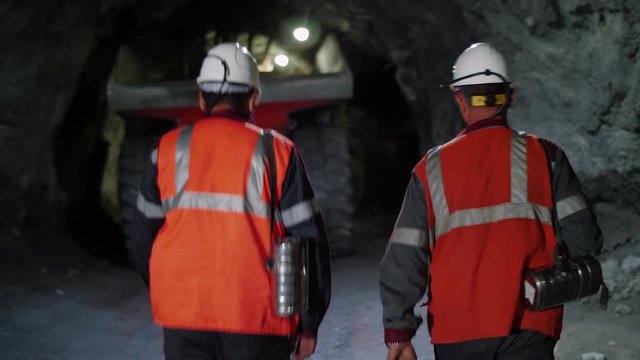 Two workers with helmets and equipment walking through a mine tunnel, in a background of heavy mining machinery.