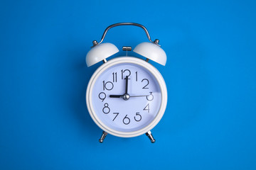 A white alarm clock on a blue background. a top view. a close-up picture representing 9 o'clock