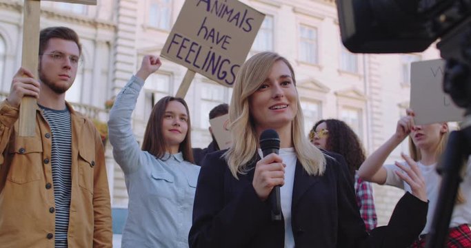 Caucasian young pretty female journalist talking in the mic in front of the camera while doing news episode from the protests of climate change activists and animals protectors.