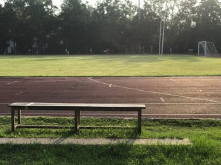 A wooden bench besides red rubber running track near the football field in the morning