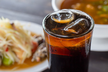 Pop water or cola in glass with ice cubes with food in the background.