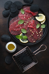 Round metal tray with carpaccio beef and black potato chips, flatlay over dark brown metal surface,...