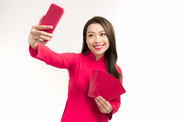 Portrait of a happy young girl dressed in ao dai taking a selfie while showing red envelope isolated over white background