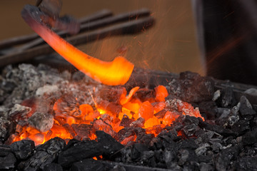 Horseshoe heating for forging. A blacksmith holds hot horseshoe with large tongs above forge. Embers glow in a iron forge. Fire, heat, coal, flying sparks.