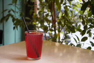 a red drink or compote in a faceted glass with a black plastic tube on a background of green trees stands on a wooden table, illuminated by natural light