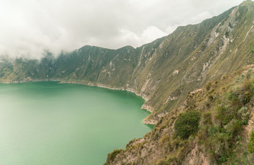 Volcano crater lake view, Quilotoa. Dramatic perspective of Quilotoa lake and volcano crater, with view of mountains, hiking path trail loop and cloudy sky from viewpoint. Shot in Ecuador. Green blue