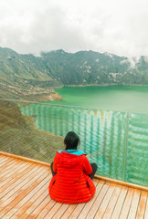 Fototapeta premium Woman at mountain volcano lake. Hiker in red looking at dramatic perspective of Quilotoa lake and crater view. Hiking loop from viewpoint. Shot in Ecuador. Green and blue. Freedom, Adventure.