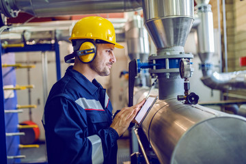 Side view of dedicated factory worker standing next to boiler and holding tablet. Worker is dressed...