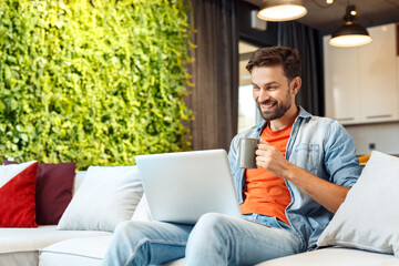 Leisure at Home. Young man sitting on sofa browsing internet on laptop checking social media cheerful drinking coffee