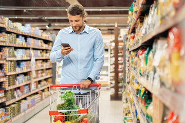 Young adult man shopping in supermarket and using smartphone