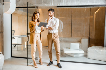 Two colleagues dressed casually meeting in the hallway, solving some working moments with digital tablet. Work in marketing agency or design studio concept