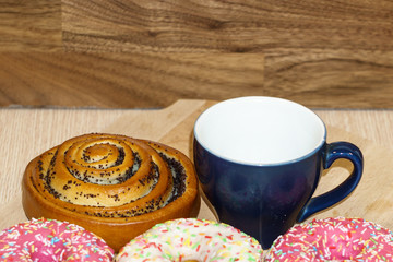 Multi-colored donuts and a bun with poppy seeds lie on a wooden table next to an empty blue cup. Top landscape with a place for text. Template for design. Spare photo.