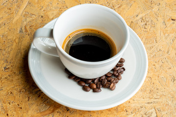 Coffee Americano cup in white saucer and roasted coffee beans under coffee cup on wood background