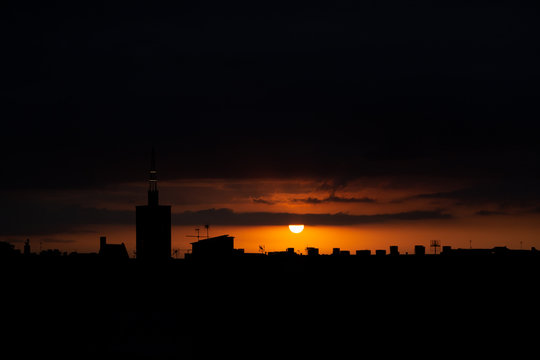 Sun appears from behind the clouds, above the city, roof top view of an old church tower. Summer sunrise or sunset. Dark orange sky color tones. Old town of Badalona, Spain
