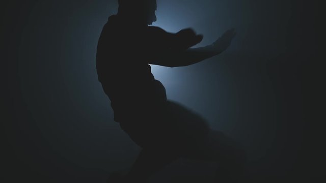 Fighter training in smoky dark gym. Close-up of male kickboxer punching with leg in jump. Fighter punching. Kickboxer training in low light gym. Silhouette on dark background in slow motion