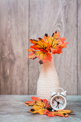 Autumn still life. A vase with orange maple leaves and a white alarm clock stand on the table. White-orange-beige color scheme. Copy space