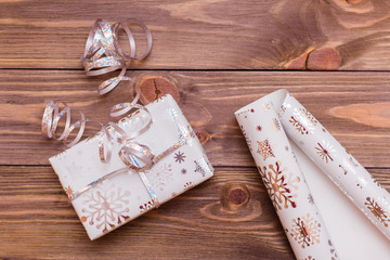 Wrapped in festive packaging box with a gift tied with silver ribbon and a roll of wrapping paper on a wooden table. Top view. Preparing for Christmas.