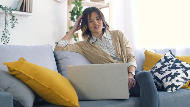 Young Woman Seat on Couch inn Modern Living Room and Using Laptop for Watching Video or Social Network. Lifestyle Concept. Happy Woman Relaxing at Home.
