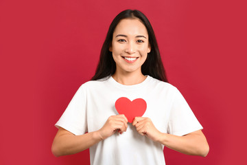 Beautiful young Asian woman with paper heart on color background