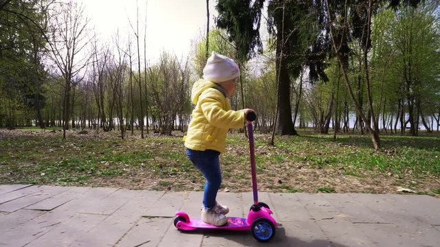 A cute little girl learns to ride a scooter in a park in early spring.