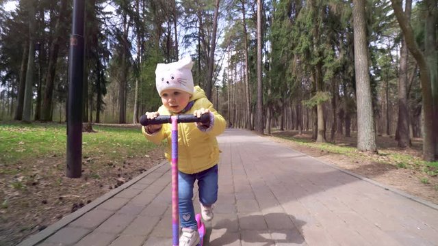 Cute little girl with great speed rides down a hill on a scooter in the park in early spring.