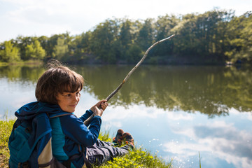 A boy with a backpack sits on the lake with a wooden stick.