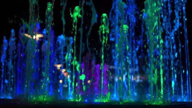 bright multi-colored fountain streams rise high above dark water surface in evening extreme close view slow motion