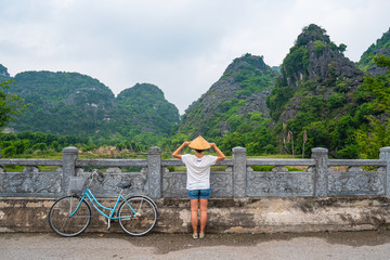 Fototapeta na wymiar Woman with traditional hat looking at unique view of Trang An Ninh Binh, Vietnam. Caucasian lady having fun on vacation riding bicycle in scenic landscape among rock pinnacles and green valleys.