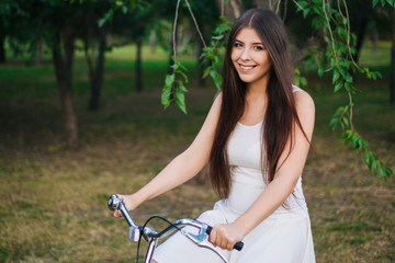 Obraz premium Young smiling girl sits on a bicycle with a basket of flowers in the evening in the park.