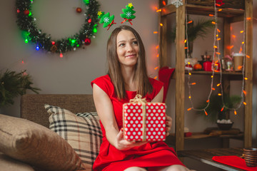 Woman in red dress sits on a sofa with a gift box in her hands on background of garland lights. Cozy Christmas mood