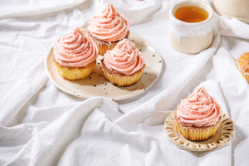 Homemade cupcakes with pink buttercream and coconut flakes served with ceramic teapot, cup of tea on white folded tablecloth.