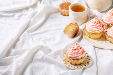 Homemade cupcakes with pink buttercream and coconut flakes served with ceramic teapot, cup of tea on white folded tablecloth.