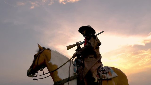 Cowboy riding across grassland with lake behind, Cowboy ranger or hunter with gun walking in forest early afternoon. Cowboy on horse riding and holding a gun in silhouette Scene.Panning around cowboy.